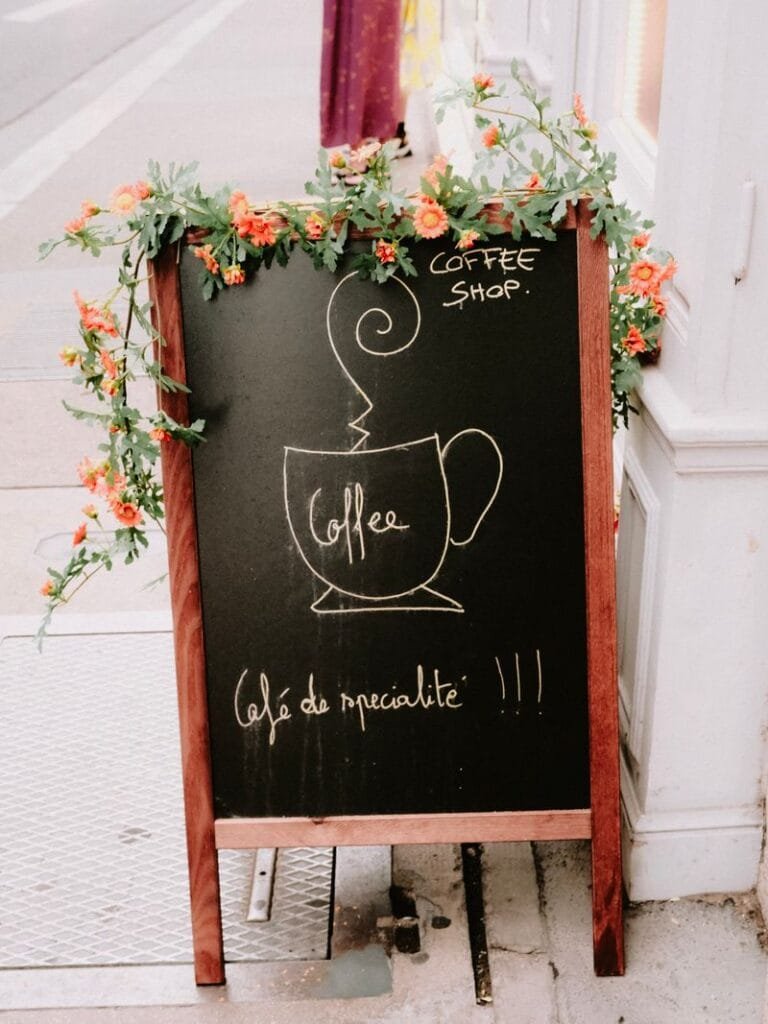 Chalkboard-style A-frame sandwich board sign advertising coffee, decorated with floral arrangements on each side